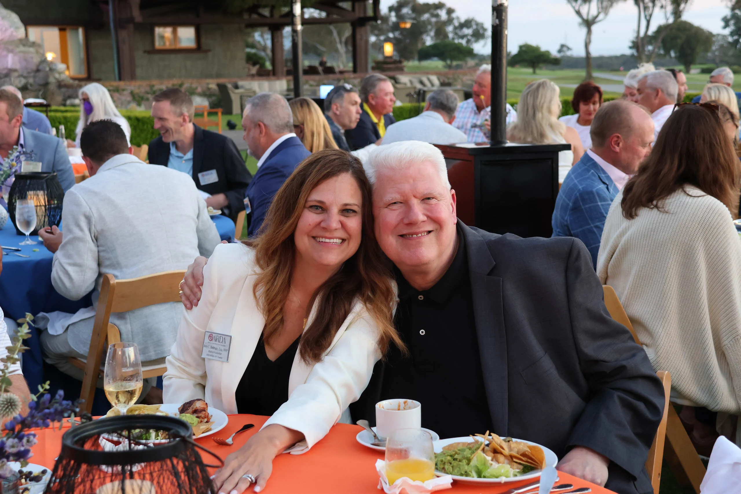 A man and woman sit at a table outdoors during a social gathering. They are smiling, with plates of food and drinks in front of them. Other people are seated at tables in the background.