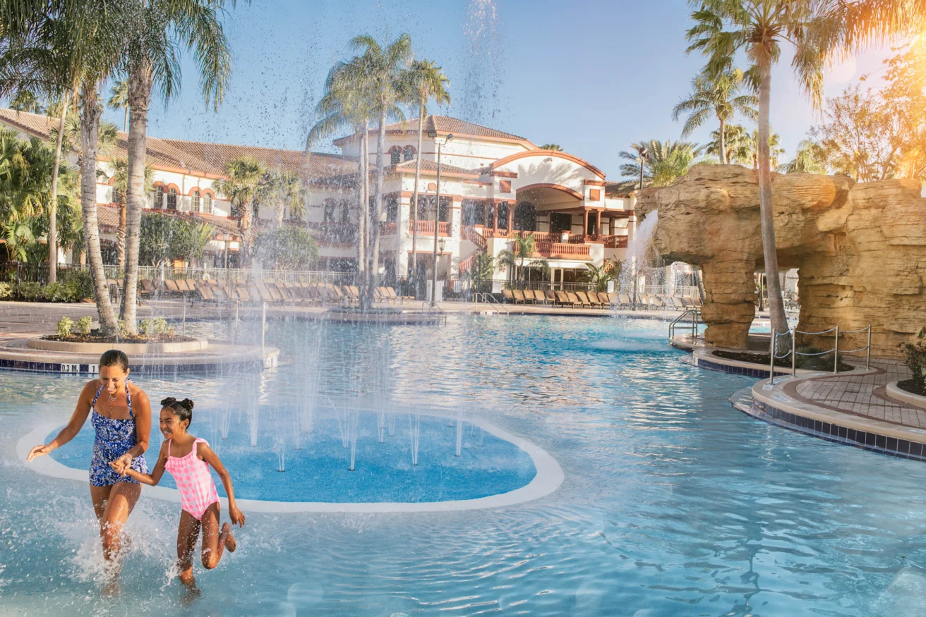 A woman and a child play in a resort's pool with fountains. Palm trees and a large building are in the background under a sunny sky.