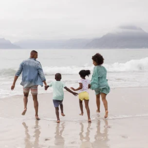 A family of four, two adults and two children, hold hands and run toward the ocean on a sandy beach, with distant mountains under a cloudy sky.