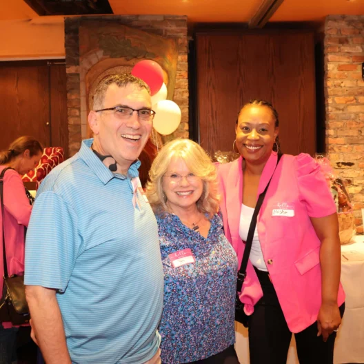 Three people smiling in a room with a brick wall and balloons in the background. Two women and one man, all wearing name tags.